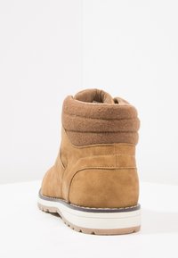 Bota de ante marrón con un acolchado en el collar, forro de tela suave y suela de goma texturizada. Presenta detalles cosidos a lo largo de las costuras.