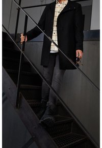 Musta takki ruudullisen paidan, harmaiden housujen ja mustien saappaiden päällä. Henkilö laskeutuu metalliporrasta ja pitää aurinkolaseja yhdessä kädessä.