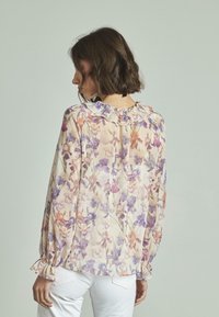 Bluse mit langen Ärmeln in Creme, mit lila und orangefarbenem Blumenmuster, Rüschenausschnitt und gerafften Bündchen. Getragen mit weißen Jeans.