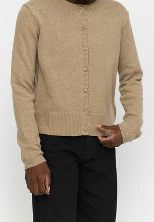 Persoon draagt een beige knoopvest en zwarte broek, met één hand die de zoom van het vest aanpast.