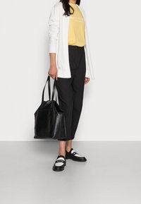 Svart läder tote-väska med dubbla handtag, bärs tillsammans med svarta byxor, vit cardigan och gul t-shirt med logotyp. Svart och vita skor.