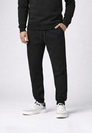 Uomo che indossa pantaloni della tuta neri con coulisse e felpa nera, in piedi con una mano in tasca, indossando sneakers bianche con dettagli neri.