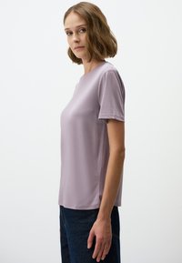 Jasnofioletowa koszulka z krótkim rękawem i okrągłym dekoltem, gładka w dotyku, o prostym kroju. Noszona z ciemnoniebieskimi dżinsami.