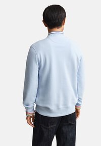 Homme portant un sweat-shirt bleu clair avec des poignets et un col rayés, vu de dos, assorti à un jean foncé.