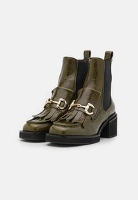 Pair of shiny olive green ankle boots with fringe detail, gold metal buckle, elastic side panels, and chunky black heels on white background.