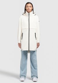 Parka bianca impermeabile con colletto alto, due tasche laterali con zip e zip frontale, abbinata a jeans a gamba larga blu chiaro.