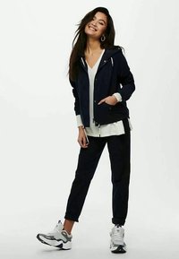 Νεαρή γυναίκα που χαμογελά, με τα χέρια στις τσέπες, φορώντας μπλε φούτερ, άσπρο μπλουζάκι, μαύρο παντελόνι και άσπρα αθλητικά, στέκεται μπροστά σε απλό φόντο.