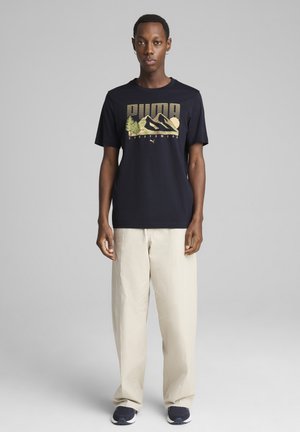 T-shirt nera in cotone con grafica di una montagna e un albero in verde e giallo. Il modello lo abbina a pantaloni larghi color crema e scarpe blu navy.