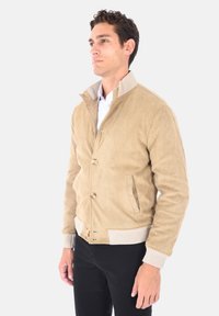 Uomo che indossa una giacca beige in suede con polsini e colletto a coste, in piedi e che guarda a sinistra contro uno sfondo bianco uniforme.
