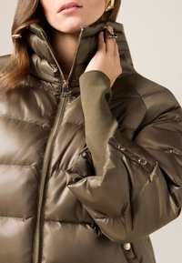 Chaqueta acolchada de color verde oliva con cuello alto, acabado brillante, cierre de cremallera y puños de punto en contraste. Sutil patrón acolchado y bolsillos laterales.