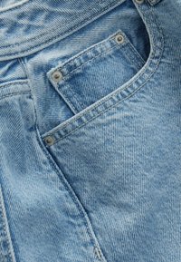 Tissu en denim bleu clair avec une poche visible, des rivets argentés et une texture subtile. Les détails de couture mettent en valeur le design de la poche et des coutures.