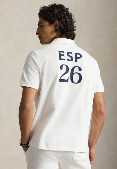 Άνδρας που φοράει λευκό πόλο με το «ESP 26» σε μπλε στο πίσω μέρος, στέκεται με το ένα χέρι στην τσέπη, κοιτάζοντας αριστερά σε απλό φόντο.