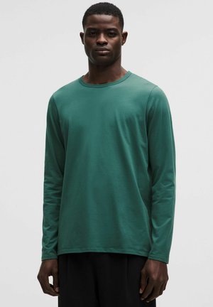 Homme debout, face à l'avant, portant une chemise verte unie à manches longues et un pantalon noir, sur un fond clair neutre.