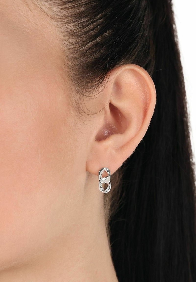 Närbild av en kvinnas öra som bär en silverörhänge med dubbel ögla, en ögla slät och en ögla beströdd med små kristaller.