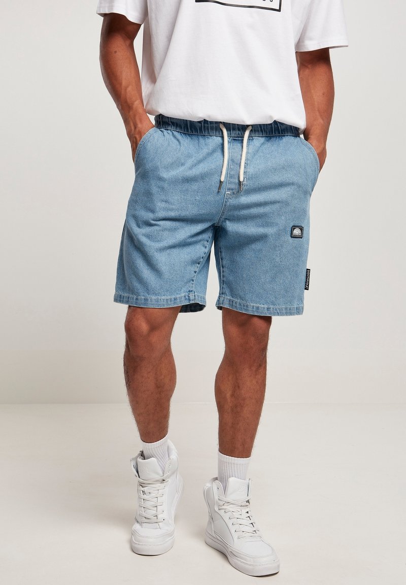Homme portant un short en jean bleu clair à taille coulissante, des baskets montantes blanches, des chaussettes blanches et une chemise blanche partiellement visible.