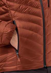 Veste brune isolée avec un design matelassé. Dotée d'une poche zippée avec une fermeture éclair noire et une languette de tirage. Matériau lisse et léger.