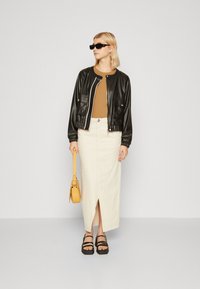 Biker in pelle nera con zip bianca, top marrone, gonna midi beige con spacco frontale, sandali platform neri e borsa gialla.