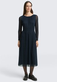 Navyblauwe midi-jurk met lange doorschijnende mouwen, textuurstof met een subtiel patroon en een aansluitende taille. Gecombineerd met zwarte plateau-schoenen.