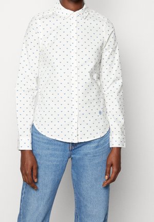 Osoba ubrana w białą koszulę na guziki z małym niebieskim wzorem motyla oraz niebieskie dżinsy, stojąca na jednolitym tle.