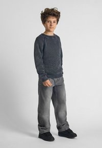 Gray knit sweater with ribbed texture, paired with light gray jeans. Black sneakers complete the outfit. The child stands with a neutral expression.