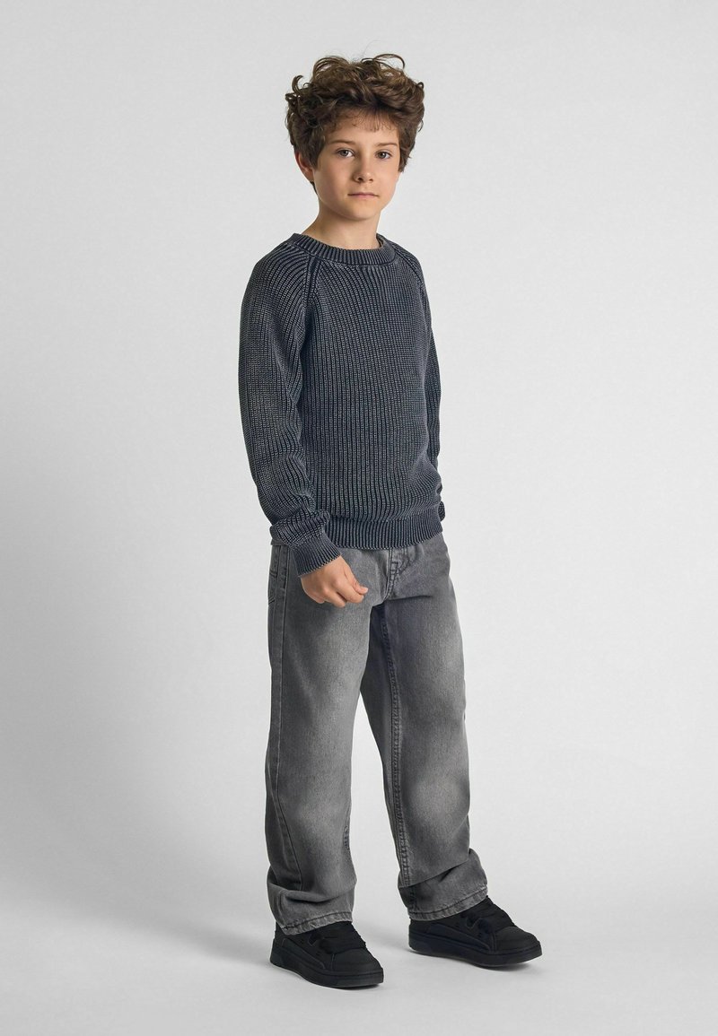 Gray knit sweater with ribbed texture, paired with light gray jeans. Black sneakers complete the outfit. The child stands with a neutral expression.