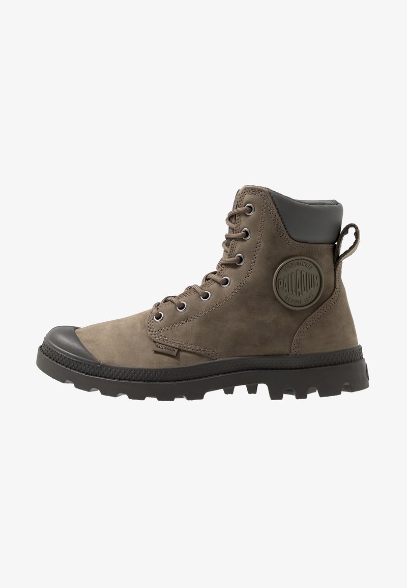 Bottine en suède marron avec embout en caoutchouc, accent gris à la cheville et semelle texturée. Présente des œillets en métal et un patch logo rond sur le côté.