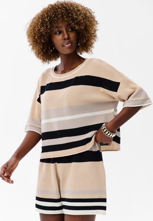 Kobieta z kręconymi włosami, nosząca beżowy, czarny i biały prążkowany dziergany top oraz pasujące szorty, pozująca z jedną ręką w kieszeni, a drugą wyciągniętą.