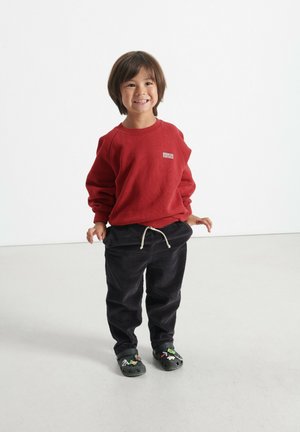 Felpa rossa con texture a coste e logo sul petto, abbinata a pantaloni in velluto nero e zoccoli neri decorati. Outfit semplice e casual.