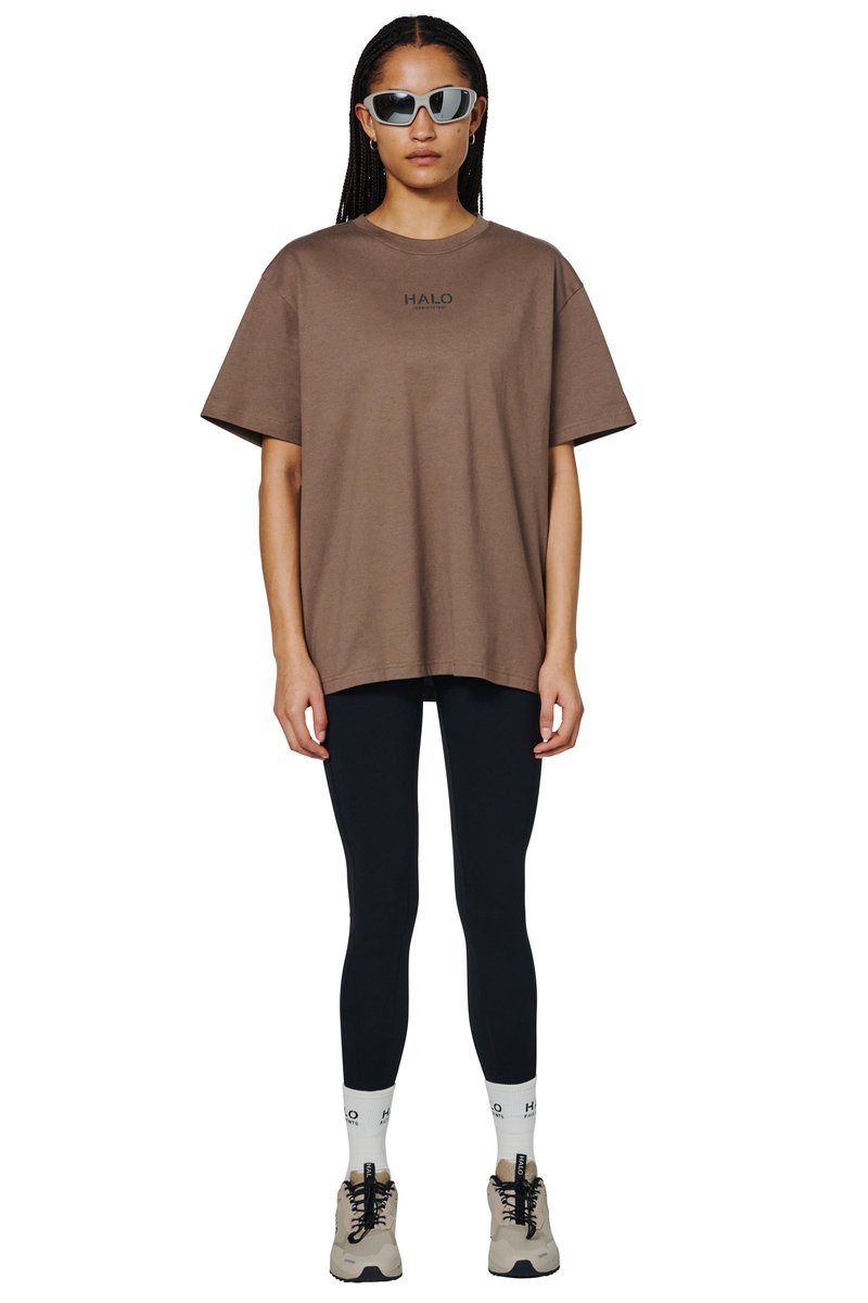 Kvinde iført oversize brun t-shirt, sorte leggings, hvide sokker, beige sneakers og sølvfarvede solbriller, stående med fronten mod kameraet på hvid baggrund.