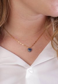 Collier en chaîne dorée avec un pendentif en forme de cœur bleu et à la surface texturée, porté sur une chemise blanche à col.