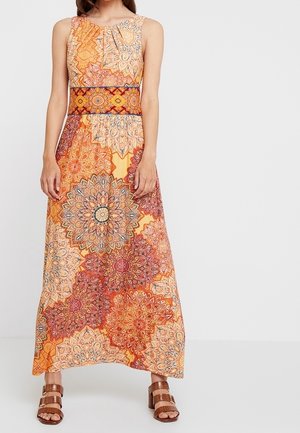 Frau trägt ein ärmelloses, knöchellanges Kleid mit orange-, rot- und beige-farbenen Mandala-Mustern und braune Sandalen mit Schnallenriemen.