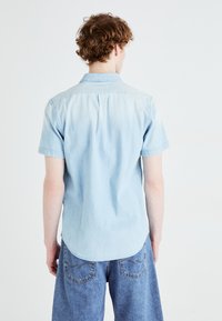 Chemise en denim bleu clair à manches courtes avec un ourlet arrondi, une poche unique et un col. Texture lisse, design décontracté.