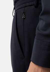 Pantalons navy en tissu lisse et texturé, dotés d'une taille élastique et d'une poche latérale avec une tirette ronde noire.