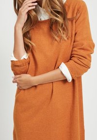 Oranžový pletený svetr s volným střihem, kulatým výstřihem a ohrnutými rukávy. Nositý přes bílou košili s límečkem. Měkká textura.