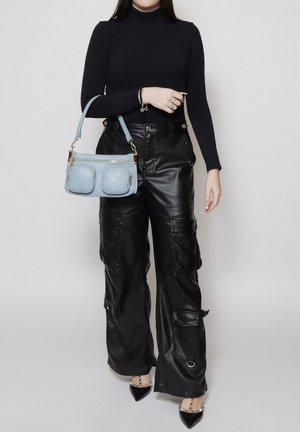 Vrouw draagt een zwarte coltrui en leren cargobroek, en houdt een lichtblauwe handtas met twee voorvakken vast.