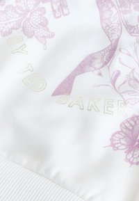 Tissu blanc avec des motifs floraux et d'oiseaux rose pâle et le texte embossé en or « BY TED BAKER » partiellement visible.