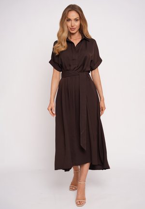 Kobieta o falujących blond włosach ubrana w ciemnobrązową, midi sukienkę z paskiem, krótkimi rękawami oraz beżowe sandały na wysokim obcasie, stojąca na białym tle.