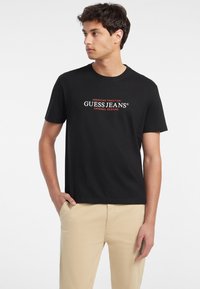Schwarzes Baumwoll-T-Shirt mit "GUESS JEANS"-Logo in weißen und roten Buchstaben, runder Ausschnitt und kurzen Ärmeln, kombiniert mit beigen Hosen.
