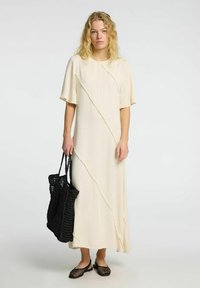 Vestido maxi crema con mangas cortas y un dobladillo asimétrico. El modelo sostiene una bolsa de tote negra de mimbre y lleva puños de malla.