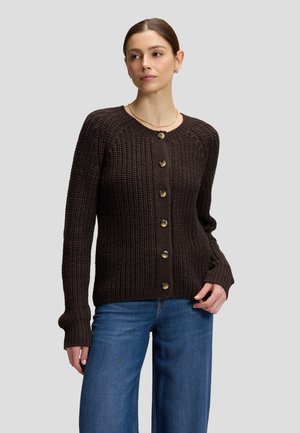 Frau trägt einen dunkelbraunen gestrickten Knopf-Cardigan und blaue Jeans und steht vor einem schlichten hellen Hintergrund.