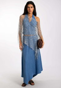 Top de mezclilla azul sin mangas con un corte asimétrico, combinado con una falda larga. Superpuesto con un wrap de malla plateada. Clutch de mimbre marrón.