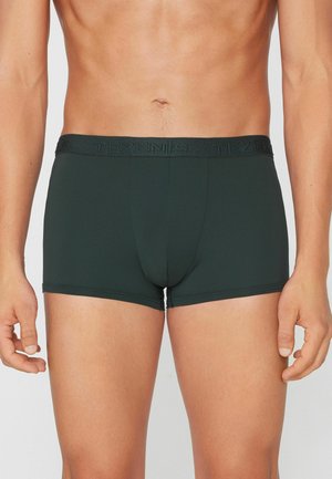 Torse masculin portant un boxer ajusté vert foncé avec une ceinture de marque, debout devant un fond clair uni.