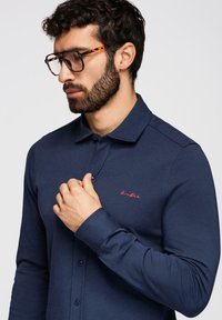 Homme aux cheveux bouclés foncés et barbe, portant des lunettes écaille de tortue et une chemise bleu marine à boutons avec un texte brodé rouge sur la poitrine.