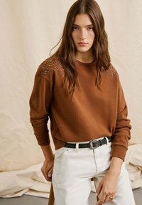 Sudadera marrón con tachuelas decorativas en los hombros, combinada con vaqueros blancos y un cinturón negro con una hebilla de plata redonda. Textura suave.