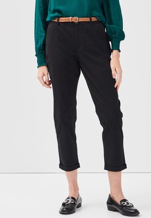 Frau trägt schwarze, gekürzte Hosen mit umgeschlagenen Bündchen, einen braunen gewebten Gürtel, eine grüne Bluse und schwarze Loafers mit silbernen Schnallen.