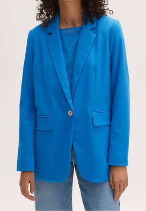 Vrouw draagt een felblauw colbert met één knoop, over een bijpassend topje en lichtblauwe spijkerbroek, met krullend haar zichtbaar aan de bovenkant.