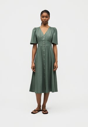 Γυναίκα που στέκεται φορώντας ένα πράσινο μίντι φόρεμα με φουσκωτά μανίκια, κουμπιά στο μπροστινό μέρος και μαύρα σαγιονάρες, σε απλό λευκό φόντο.