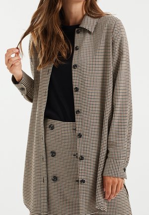 Femme portant un manteau et une jupe assortie à motif pied-de-poule beige et noir sur un haut noir, avec des boutons sur le devant des deux vêtements.