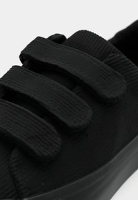 Svart joggesko med teksturert materiale og tre borrelåsremmer. Rund tå og gummisåle. Nærbilde som fremhever detaljer på remmene.