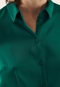 Camicia teal con bottoni, realizzata in un tessuto liscio e setoso, dotata di un colletto classico e cinque bottoni coordinati sul davanti.